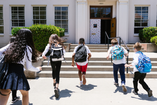 Five children wearing backpacks run towards a school.
