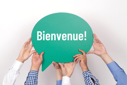Plusieurs mains brandissent une pancarte en forme de bulle. Sur le panneau, il est écrit « Bienvenue! »