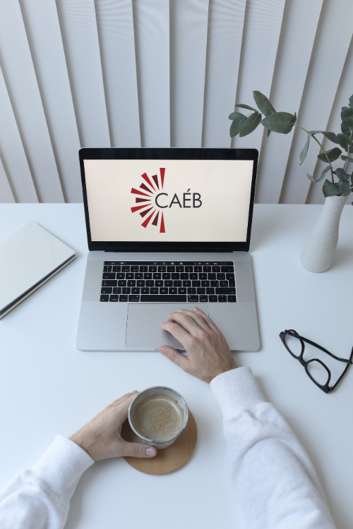 Un ordinateur portable est posé ouvert sur une surface blanche et une paire de lunettes est posée à côté. Une paire de mains est posée devant le clavier, l'une sur le trackpad et l'autre autour d'une tasse.