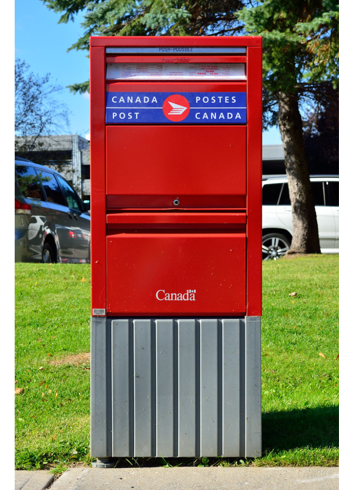 Une boîte aux lettres de Postes Canada est posée dans l'herbe, au bord d'un trottoir.