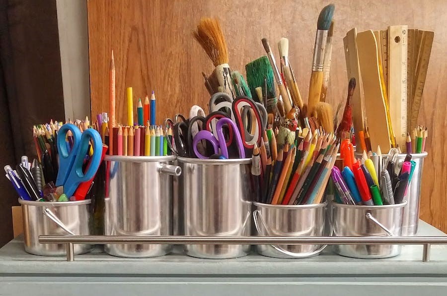 Many tins containing art supplies such as scissors, paintbrushes, rulers, pens and coloured pencils sit on a shelf.