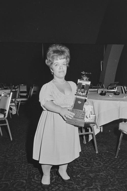 Judy-Lynn del Rey is holding an award while standing in a banquet hall.
