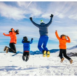A family of 4 dressed in winter clothing jump up in the air. They are outdoors in the snow and the mountains are in the distance.