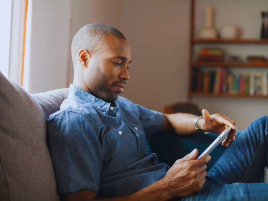 A man sits on a couch and reads on a tablet device.