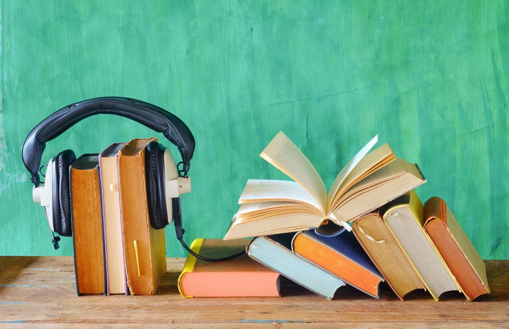 A pile of books on a wood surface and three books lined upright beside them with over-ear headphones over them.