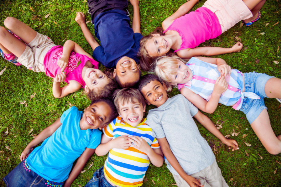 Six enfants sont étendus dans l'herbe en cercle, leurs têtes se touchant, souriant à la caméra.