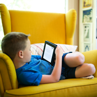 A young boy curls up on a chair while reading a book on a tablet.