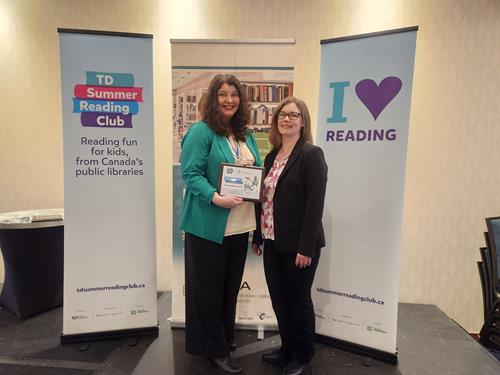 Two women, Andrea Siemens and Rachel Breau stand in front of TDSRC banners. Andrea is holding a plaque.