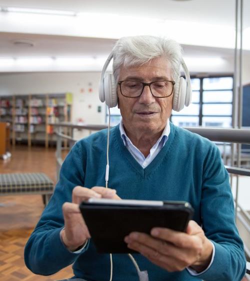 Un homme âgé porte des écouteurs et tient une tablette alors qu'il est assis dans une bibliothèque.