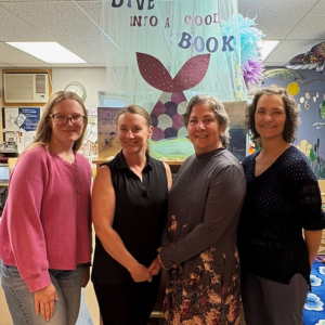 Four women stand in front of library shelves smiling at the camera