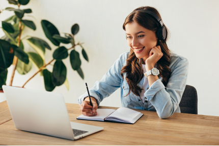 A woman sitting at a table with headphones on writes in a notepad and smiles at a laptop open in front of her.