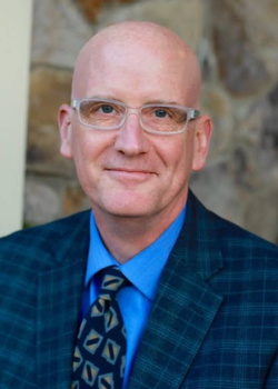 Dan Willingham, a bald man wearing glasses and a blue shirt and suit jacket smiles at the camera.