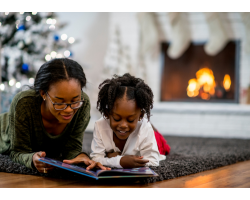 A young woman and a little girl read a book beside a Christmas tree. There is a fireplace in the background with stockings hung above.