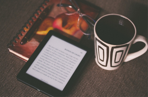 Une liseuse est posée sur un carnet, près d’une paire de lunettes et d’une tasse de café