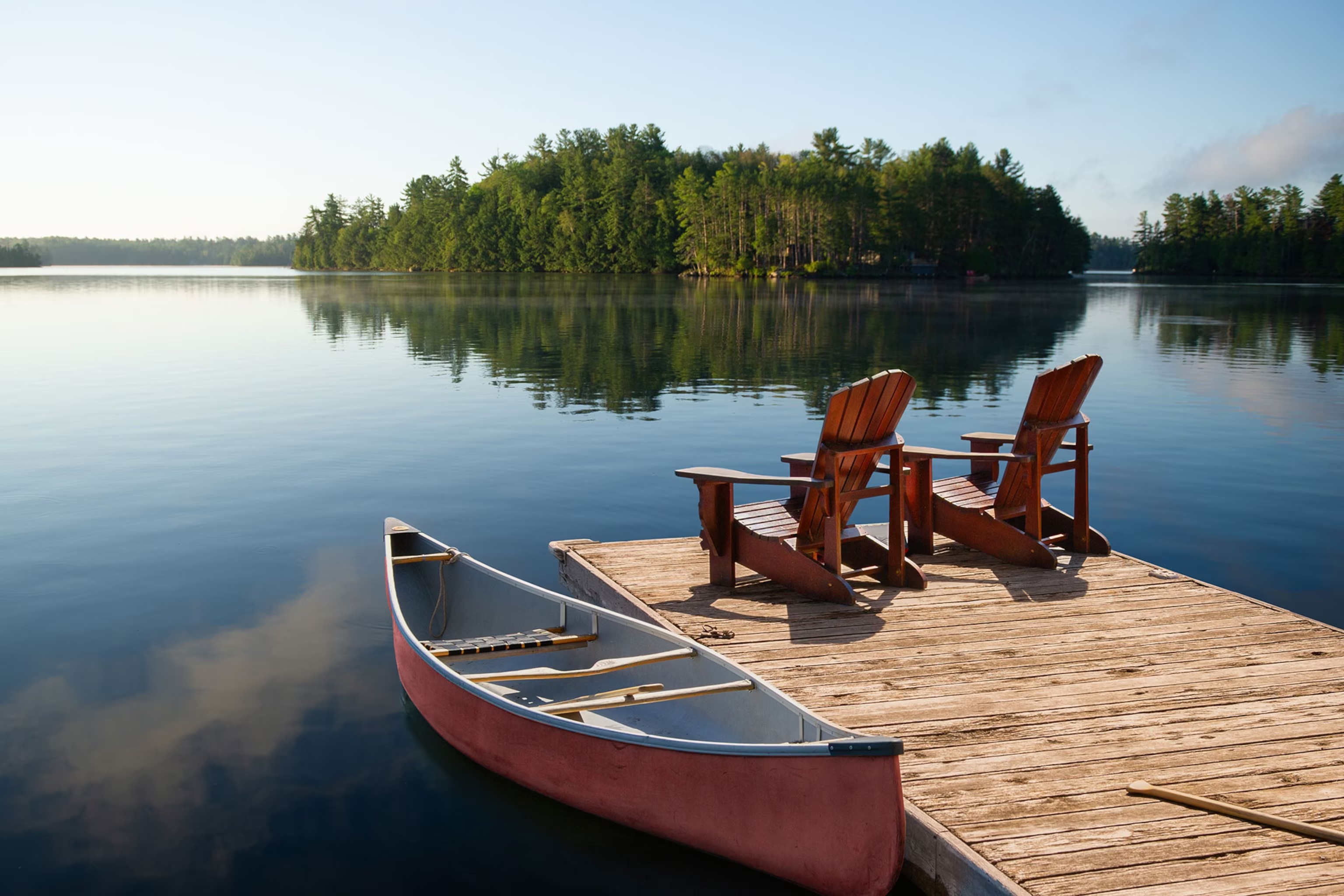 Un paysage de lac avec un canot et deux chaises Adirondack sur un quai.