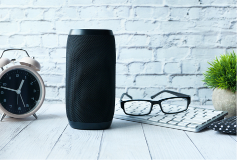 A smart speaker on a desk with an alarm clock, a pair of glasses and a computer keyboard.