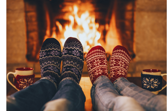 Deux personnes visibles à partir des genoux se réchauffent les pieds devant le feu d'une cheminée. Ils portent des chaussettes festives et ont des tasses festives à côté d'eux.