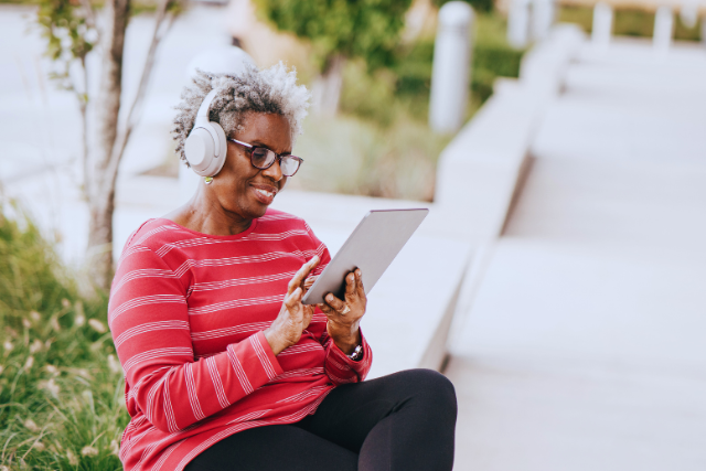 Une personne portant des lunettes et des écouteurs est assise à l'extérieur sur un banc et tient une tablette.