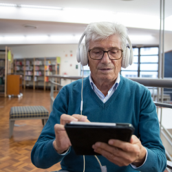 Un homme âgé est assis dans une bibliothèque, écoutant au casque tout en tenant une tablette.