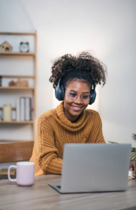 A woman wearing headphones sits at a table and smiles at a laptop.